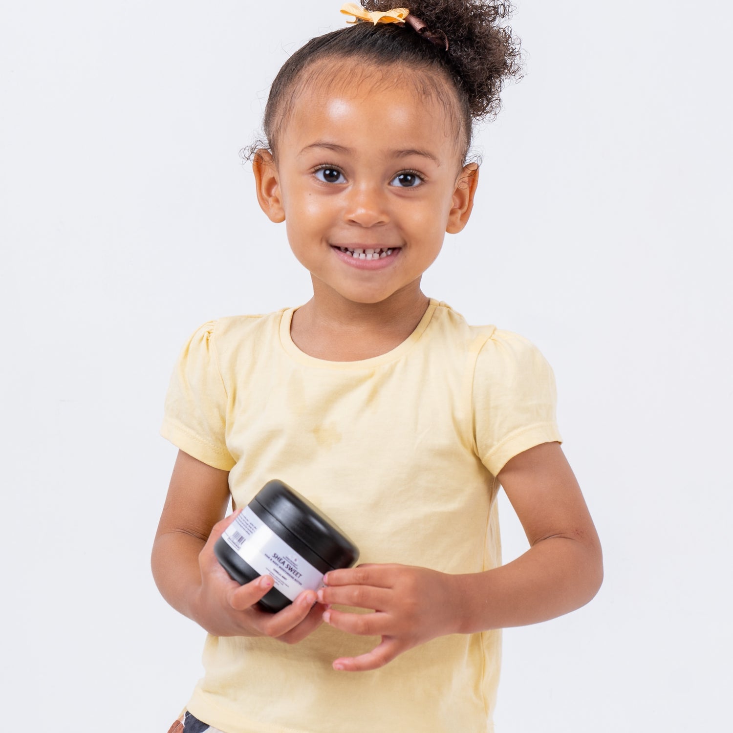A curly haired, smiling girl holding a black tub of vanilla mint shea sweet in both hands