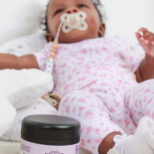 Baby in pink floral outfit with a pacifier, next to a jar of Official True Beauty Shea sweet product.
