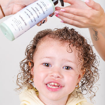 Close-up of child with wet curly hair smiling as Butter Curls Vanilla Mint leave-in conditioner is dispensed into an adult’s hand.