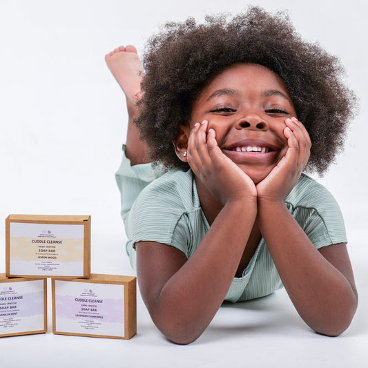 Smiling Child with a green top lying on a white surface with Cuddle Cleanse soap bars in the foreground